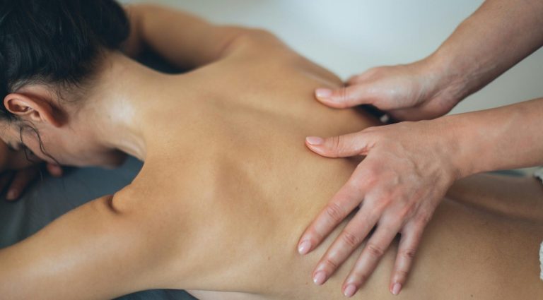 Dunkelhaarige Frau liegt auf der Massageliege. Masseurin drückt bewusst auf die Triggerpunkte am Rücken.